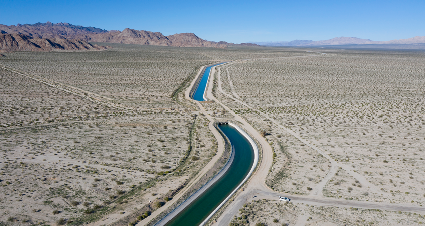 A view of a canal carrying water through a large, arid swath of land with mountains in the background.