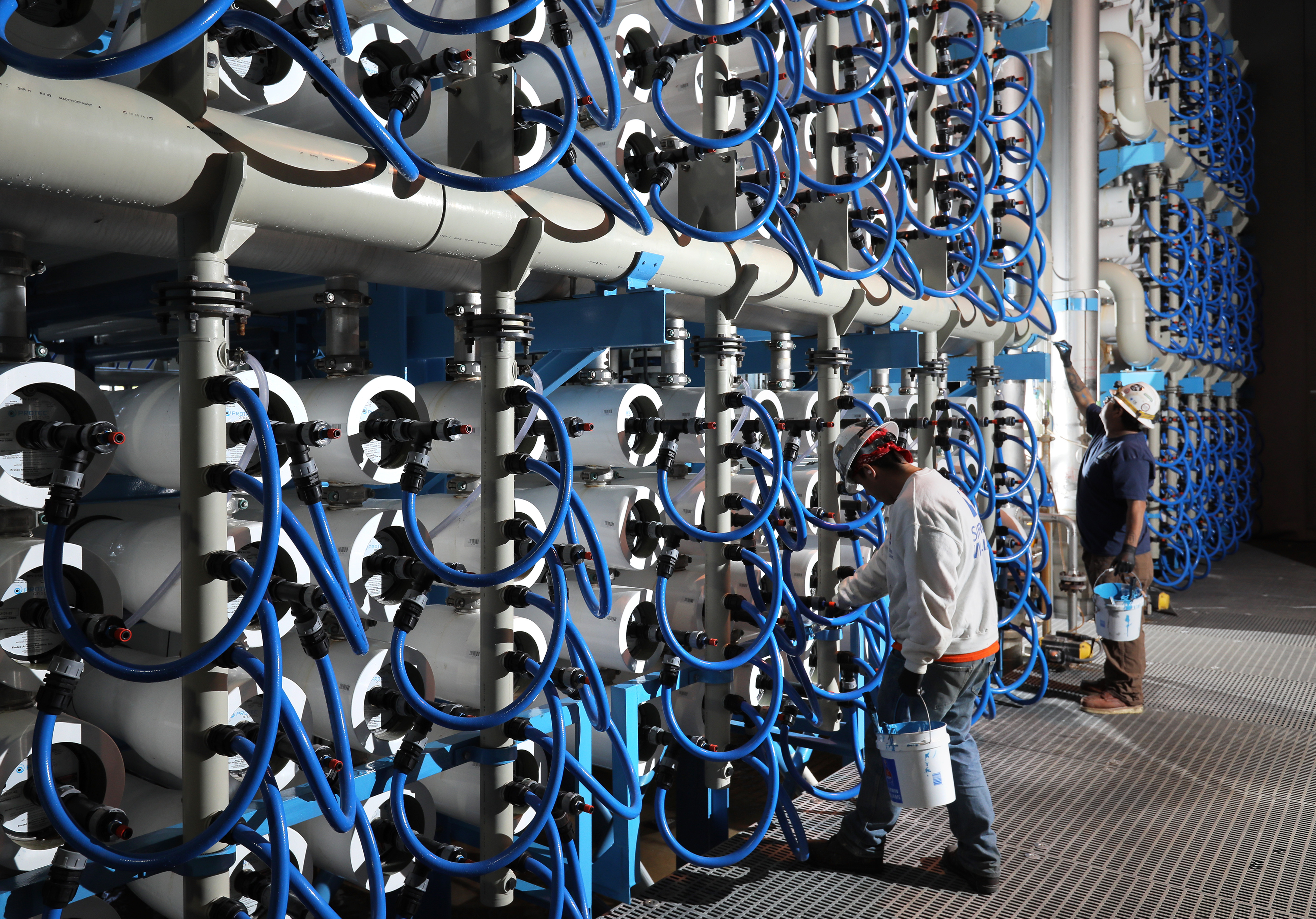 Employees at the Carlsbad Desalination Plant stand in a large room with hundreds of reverse osmosis filters and connected pipes and machinery.